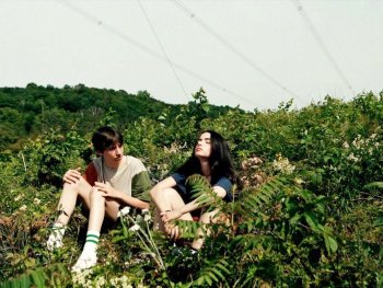 Copyright: Filmverleih Zwei junge Personen sitzen nebeneinander in einer üppig bewachsenen Wiese mit Sträuchern und Blumen unter freiem Himmel.