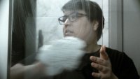 Person mit Brille berührt eine Glasscheibe, durch die ihr Gesicht verzerrt und spiegelnd zu sehen ist.