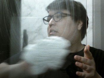 Copyright: Denis Côté Person mit Brille berührt eine Glasscheibe, durch die ihr Gesicht verzerrt und spiegelnd zu sehen ist.