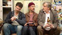 Drei Männer sitzen nebeneinander in einer Umkleidekabine mit Tennis-Aufklebern an den Spinden, einer schaut auf die Uhr, einer hält ein Tablet und einer eine Bierflasche.