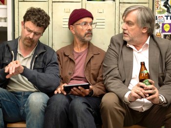 Copyright: Studiocanal GmbH Drei Männer sitzen nebeneinander in einer Umkleidekabine mit Tennis-Aufklebern an den Spinden, einer schaut auf die Uhr, einer hält ein Tablet und einer eine Bierflasche.