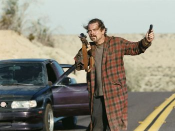 Copyright: Warner Bros. Entertainment GmbH Ein Mann in kariertem Morgenmantel steht mit Gewehr neben einem Auto auf einer Landstraße und winkt mit erhobener Hand.