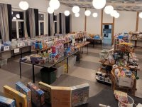 Mehrere Tische mit Spielzeug, Spielen und Kuscheltieren stehen in einem Saal mit runden Hängeleuchten für einen Second-Hand-Basar bereit.