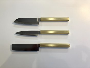Drei japanische Küchenmesser mit unterschiedlich geformten Klingen und goldfarbenen Griffen liegen parallel nebeneinander auf einer weißen Fläche.
