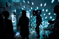 Eine Discokugel wirft Lichtpunkte auf Wände mit Fotografien, während Personen als Silhouetten in dem dunklen Ausstellungsraum stehen.