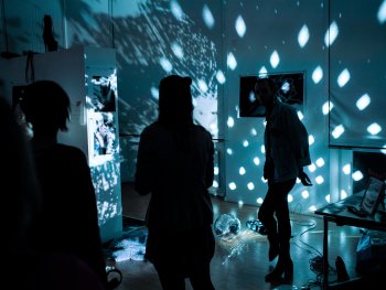 Eine Discokugel wirft Lichtpunkte auf Wände mit Fotografien, während Personen als Silhouetten in dem dunklen Ausstellungsraum stehen.