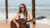 Lächelnde Person mit Sonnenbrille spielt Akustikgitarre am Strand vor Wellen und zwei Holzpfählen.