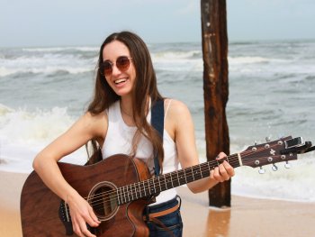 Lächelnde Person mit Sonnenbrille spielt Akustikgitarre am Strand vor Wellen und zwei Holzpfählen.