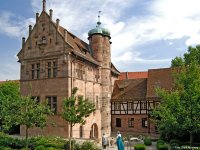 Historisches Sandsteingebäude mit rundem Turm und angrenzendem Fachwerkhaus, davor ein begrünter Innenhof mit Bäumen und Sitzgelegenheiten.