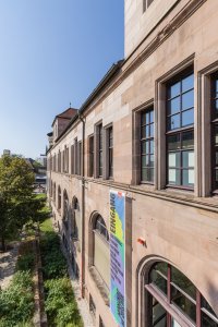 Historisches Sandsteingebäude mit großen Sprossenfenstern, an dem ein Banner mit der Aufschrift „Eingang“ zum Kulturzentrum hängt.