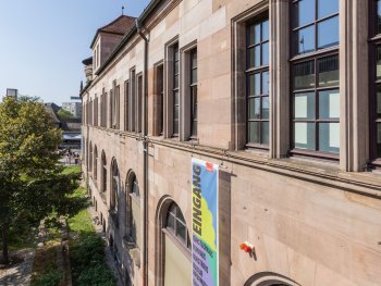 Historisches Sandsteingebäude mit großen Sprossenfenstern, an dem ein Banner mit der Aufschrift „Eingang“ zum Kulturzentrum hängt.