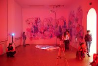 In rotes Licht getauchter Ausstellungsraum mit großflächigen abstrakten Wandmalereien, in dem Personen mit Masken zwischen Kameras und Stativen die Kunstinstallation betrachten.