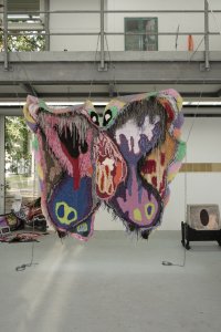 Eine großformatige, bunte Textilskulptur in Schmetterlingsform hängt an Ketten von einem Gerüst in einem Ausstellungsraum.