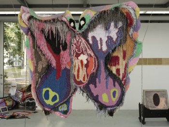 Eine großformatige, bunte Textilskulptur in Schmetterlingsform hängt an Ketten von einem Gerüst in einem Ausstellungsraum.