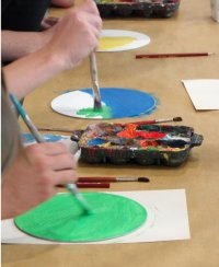 Mehrere Personen malen mit Pinseln bunte Kreise auf Papier, während in der Bildmitte eine Palette mit Farbresten liegt.