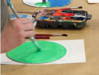 Mehrere Personen malen mit Pinseln bunte Kreise auf Papier, während in der Bildmitte eine Palette mit Farbresten liegt.