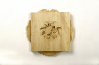 Quadratische Holzskulptur mit einer blütenartig geschnitzten Vertiefung in der Mitte, montiert auf einer runden Holzscheibe mit unregelmäßigem Rand an einer weißen Wand.