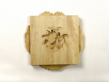 Quadratische Holzskulptur mit einer blütenartig geschnitzten Vertiefung in der Mitte, montiert auf einer runden Holzscheibe mit unregelmäßigem Rand an einer weißen Wand.