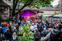 Eine große Menschenmenge verfolgt einen Konzertauftritt einer Band auf einer überdachten Open-Air-Bühne im "KulturGarten" vor einer alten Sandsteinmauer.