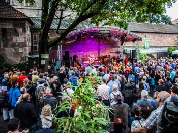Eine große Menschenmenge verfolgt einen Konzertauftritt einer Band auf einer überdachten Open-Air-Bühne im "KulturGarten" vor einer alten Sandsteinmauer.
