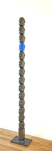 Schmale Turmskulptur aus vielen übereinander gestapelten, identischen bronzefarbenen Gesichtsmasken mit einer einzelnen blauen Maske dazwischen, auf einem Metallsockel stehend.