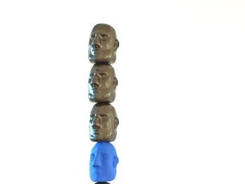 Schmale Turmskulptur aus vielen übereinander gestapelten, identischen bronzefarbenen Gesichtsmasken mit einer einzelnen blauen Maske dazwischen, auf einem Metallsockel stehend.