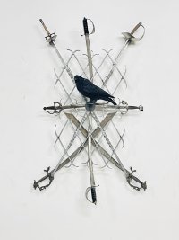 Mehrere gekreuzt angeordnete Schwerter und Säbel in einem Metallgitter steckend, das an einer weißen Wand befestigt ist, in deren Mitte ein ausgestopfter schwarzer Vogel sitzt.