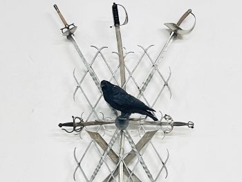 Mehrere gekreuzt angeordnete Schwerter und Säbel in einem Metallgitter steckend, das an einer weißen Wand befestigt ist, in deren Mitte ein ausgestopfter schwarzer Vogel sitzt.