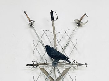 Foto: Hubertus Hess, VNP
Mehrere gekreuzt angeordnete Schwerter und Säbel in einem Metallgitter steckend, das an einer weißen Wand befestigt ist, in deren Mitte ein ausgestopfter schwarzer Vogel sitzt.