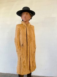 Lebensgroße Holzskulptur einer Person mit schwarzem Hut und bemaltem Gesicht, der Körper mit unbehandeltem, grob strukturiertem Holz als Mantel gestaltet.