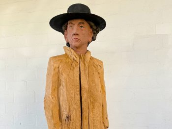 Lebensgroße Holzskulptur einer Person mit schwarzem Hut und bemaltem Gesicht, der Körper mit unbehandeltem, grob strukturiertem Holz als Mantel gestaltet.