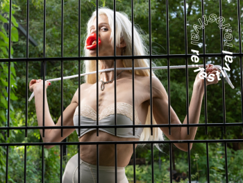 Person mit langen blonden Haaren, rotem Objekt vor dem Mund und einer Kordel in den Händen steht hinter einem schwarzen Metallzaun, im Hintergrund Grünpflanzen, mit dem Schriftzug „re:festival“.