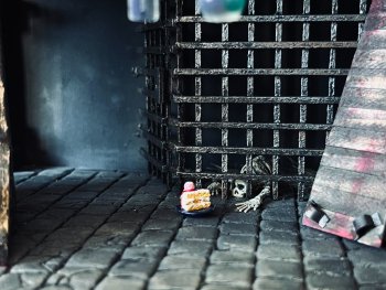 Miniaturmodell einer Gefängniszelle mit Gitterstäben, hinter denen ein Skelett liegt, während davor ein Kuchenstück auf dem Steinboden steht.