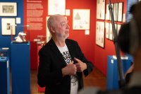 Ein Mann mit weißem Bart spricht gestikulierend vor einer Ausstellungswand mit gerahmten Zeichnungen und Vitrinen mit Skulpturen.