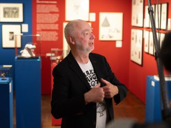 Ein Mann mit weißem Bart spricht gestikulierend vor einer Ausstellungswand mit gerahmten Zeichnungen und Vitrinen mit Skulpturen.