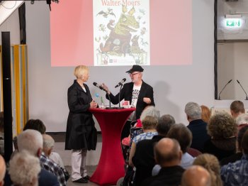 Eine Frau und ein Mann stehen an einem Bartisch und unterhalten sich. Im Hintergrund ist das Plakat der Walter-Moers-Ausstellung zu sehen. Im Vordergrund sitzt Publikum.