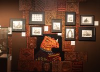 Ausstellungswand mit gerahmten Fantasy-Zeichnungen und einem großen Wandbild eines echsenartigen Wesens vor ornamental gemusterter Tapete.