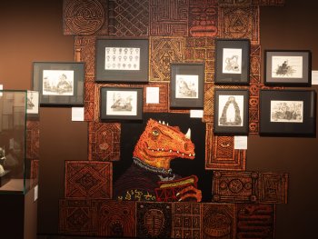 Ausstellungswand mit gerahmten Fantasy-Zeichnungen und einem großen Wandbild eines echsenartigen Wesens vor ornamental gemusterter Tapete.