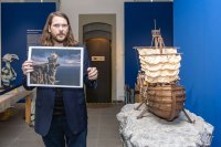 Eine Person hält ein gerahmtes Bild eines Segelschiffs auf einer Klippe neben einem detailreichen Schiffsmodell auf einem Felsen in einer Ausstellung.