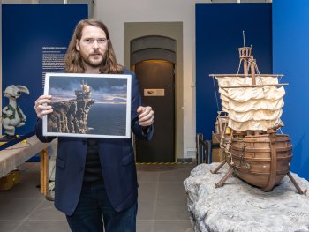 Eine Person hält ein gerahmtes Bild eines Segelschiffs auf einer Klippe neben einem detailreichen Schiffsmodell auf einem Felsen in einer Ausstellung.