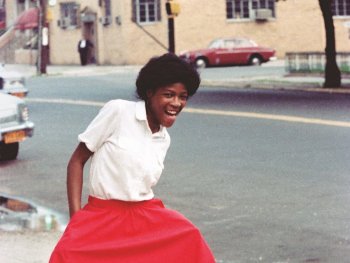 Foto: Jamel Shabazz
Lachende Frau in weißer Bluse und rotem Rock posiert ausgelassen auf einem Gehweg an einer Straße mit parkenden Autos.