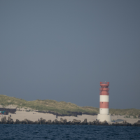Rot-weiß gestreifter Leuchtturm auf einer Insel mit Sanddünen, davor Wellenbrecher aus Steinen und das Meer.