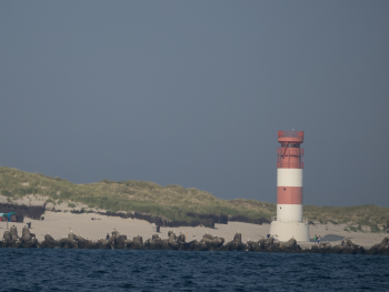 Rot-weiß gestreifter Leuchtturm auf einer Insel mit Sanddünen, davor Wellenbrecher aus Steinen und das Meer.