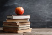 Ein roter Apfel liegt auf einem Stapel alter Bücher vor einer verwischten Tafel im Hintergrund.