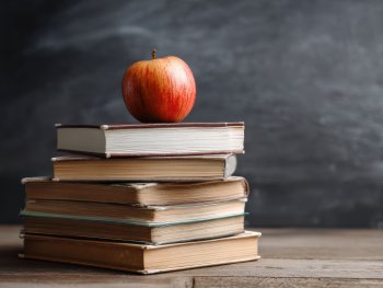 Copyright: AdobeStock Ein roter Apfel liegt auf einem Stapel alter Bücher vor einer verwischten Tafel im Hintergrund.