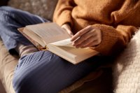 Person in Strickpullover sitzt im Schneidersitz auf einem Sofa und blättert in einem aufgeschlagenen Buch, dass auf ihrem Schoß liegt.