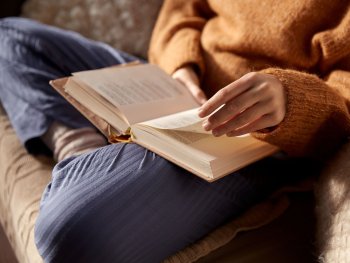 Copyright: AdobeStock Person in Strickpullover sitzt im Schneidersitz auf einem Sofa und blättert in einem aufgeschlagenen Buch, dass auf ihrem Schoß liegt.