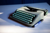 Alte graue Reiseschreibmaschine mit türkisen Tasten steht neben einem blauen Stift auf einem weißen Tisch.