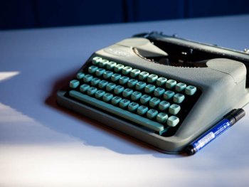 Eine graue Reiseschreibmaschine mit türkisen Tasten steht auf einem Tisch neben einem blauen Stift.