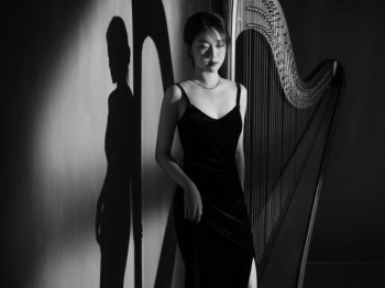 Copyright: Boija Yu Schwarz-Weiß-Porträt einer Frau in dunklem Kleid, die neben einer Harfe steht, mit deutlichem Schattenwurf an der Wand.